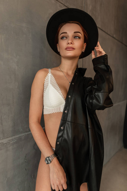 Красива сексуальна жіноча модель з струнким тілом в модній сексуальній білій нижній білизні з чорною сорочкою регулює її чорний капелюх і стоїть біля сірої стіни
 - Фото, зображення