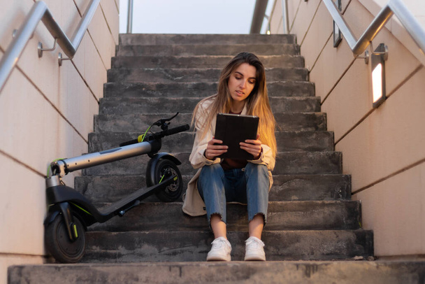 Woman sitting on steps using a tablet with an electric scooter resting on her side. With night light and spotlight - Foto, Imagen
