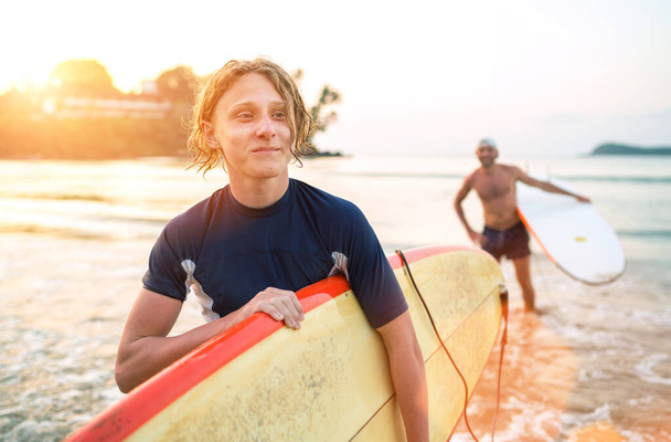 Portrait of a teen boy with a surfboard after surfing with his father. They are smiling and walking out of the water. Family active vacation concept. - Photo, image