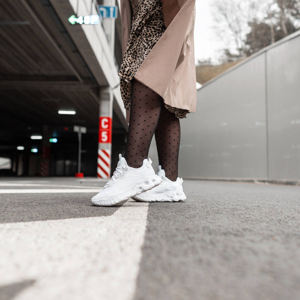 Стильна жінка в моді біле взуття з колготками, леопардовим платтям, пальто гуляє по асфальту в місті. жіночі спортивні кросівки в жіночих ногах, крупним планом
 - Фото, зображення