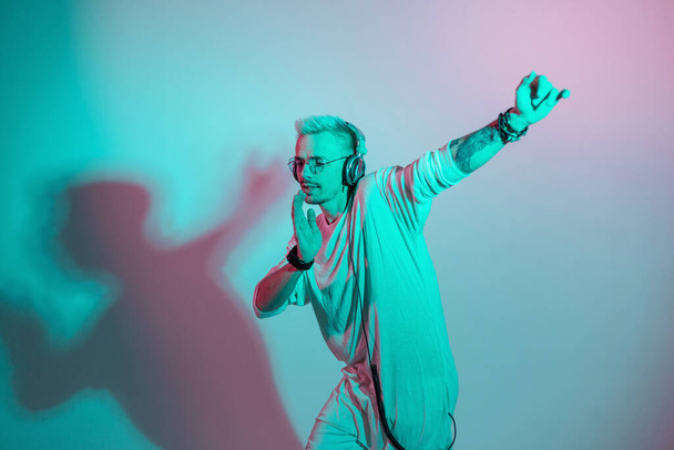 Der Hipster hört Musik und tanzt auf einem farbenfrohen rosa und blauen Hintergrund. Modemann mit Kopfhörern bewegt sich im Tanzrhythmus im Studio - Foto, Bild