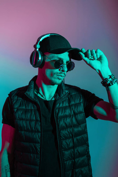 Brutal handsome man with sunglasses in fashionable black outwear straightens his cap in the studio with creative colored light - Foto, afbeelding