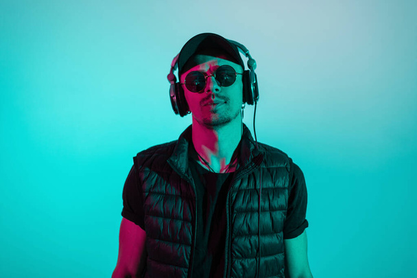 Stylish trendy man hipster in black cap, sunglasses and headphones in black fashion clothes in studio with blue and pink light color - 写真・画像