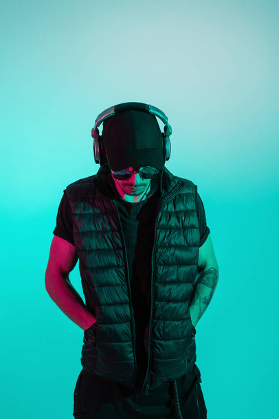 Cool man in black stylish clothes with sunglasses, fashion black cap, vest and t-shirts listening to music in headphones. Creative male portrait of trendy hipster guy in colorful background - Photo, Image