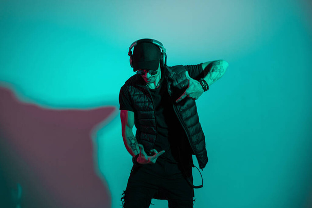 Κομψός χορευτής άντρας με γυαλιά ηλίου και καπέλο σε μοντέρνα μαύρα ρούχα που χορεύει και ακούει μουσική σε ακουστικά στο στούντιο με δημιουργικό χρωματιστό φως - Φωτογραφία, εικόνα