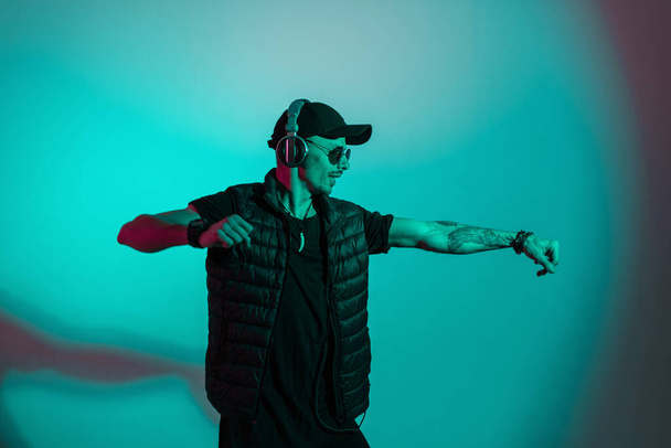 Fashion man dancer in black stylish clothes with cap and sunglasses dances in creative studio with color cyan light - Valokuva, kuva