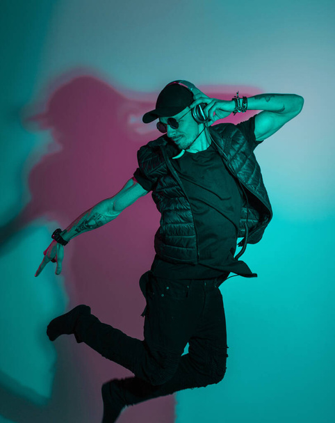 Stylish handsome male dancer in fashion black clothes with sunglasses, a cap and headphones listens to music and dances in the studio with creative color pink blue light - Photo, Image
