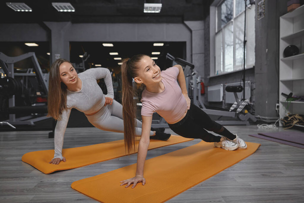 Девочка-подросток делает упражнения на доске в тренажерном зале, тренируется с личным тренером - Фото, изображение