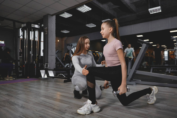 Teen girl doing lunges with weights, working out with personal trainer at the gym - Photo, Image