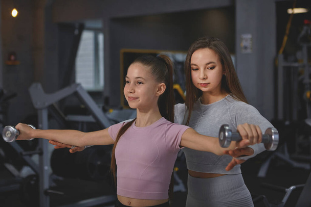 Professional sportswoman supervising teenage girl during gym workout, doing dumbbells exercise - Foto, Imagen