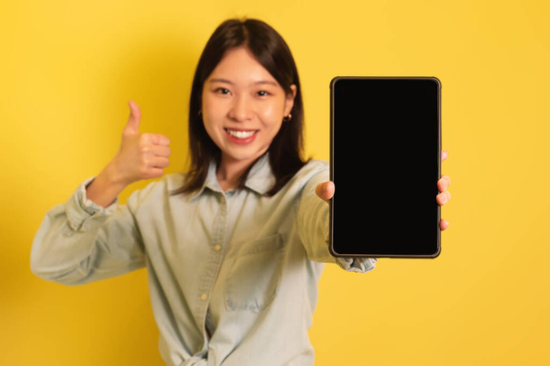 Hübsche Asiatin demonstriert digitales Tablet mit leerem Bildschirm, zeigt Daumen nach oben Geste, Attrappe für Website oder App - Foto, Bild