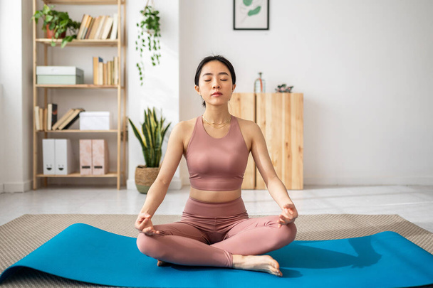 Asian woman meditating in lotus position while doing yoga at home. - 写真・画像