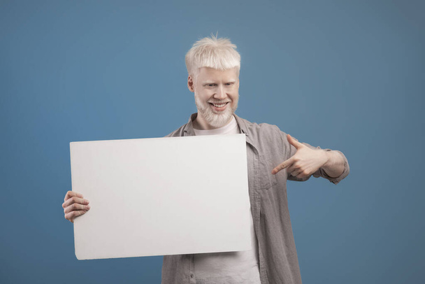 Place for your ad. Excited albino guy holding empty blank board and pointing at it, posing on blue background - Foto, Imagen