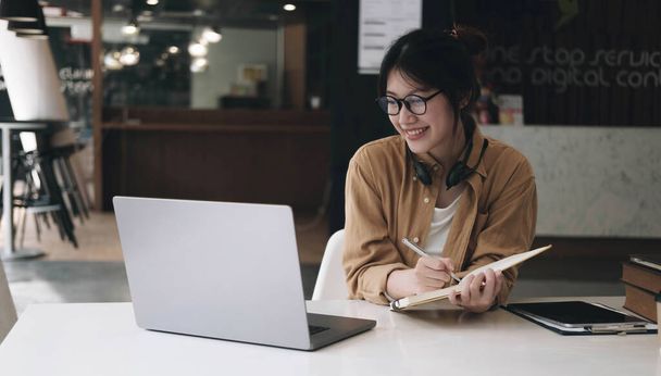 Boldog ázsiai üzletasszony visel headset, hogy jegyzetek beszélni konferenciahívás csinál video chat laptop az irodában, mosolygó kínai támogató szolgálat ügynök írni információkat olvasni custome - Fotó, kép