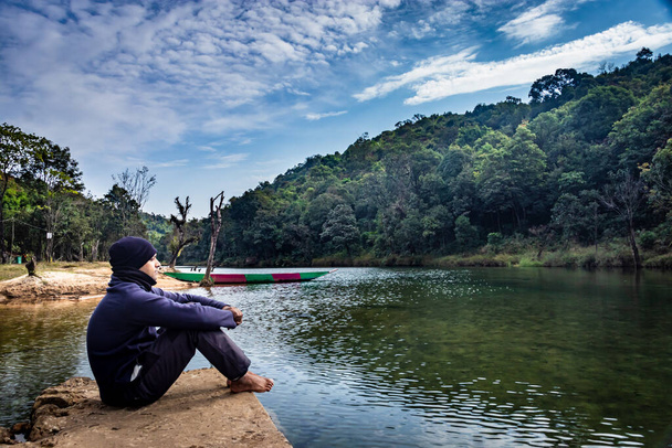 Sakin nehrin kıyısında, yoğun yeşil ormanlarla çevrili ve mavi gökyüzü ile kaplı yalnız bir adam oturuyor. - Fotoğraf, Görsel
