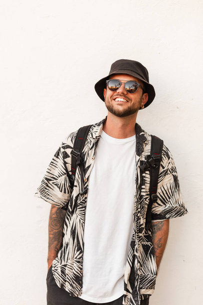 Handsome hipster man with smile in summer fashion clothes with sunglasses, hat and shirt with backpack travel in city - Фото, изображение