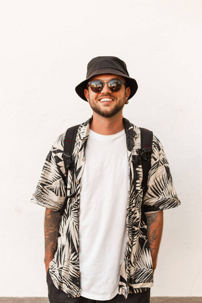 Happy young hipster man with smile in fashion summer clothes with bucket hat, shirt and sunglasses with backpack stands near white wall on the street - Фото, изображение