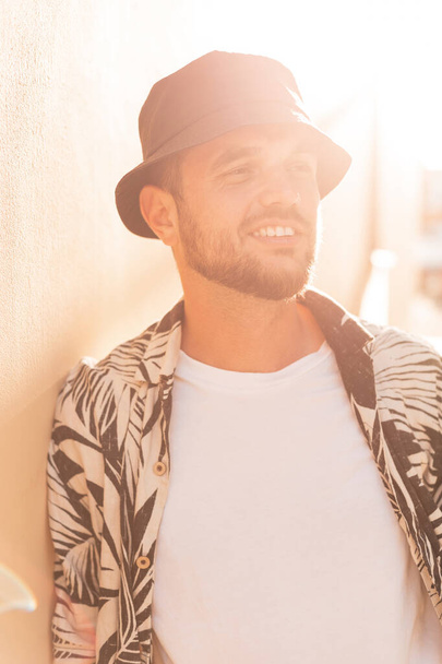 Ευτυχισμένος όμορφος hipster άντρας με χαμόγελο στη μόδα πουκάμισο με φοίνικες εκτύπωσης και μαύρο καπέλο κουβά στο ηλιοβασίλεμα - Φωτογραφία, εικόνα