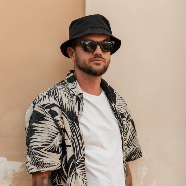 Handsome stylish hipster man with beard, sunglasses and bucket hat in fashion summer outwear with beach shirt near the wall on the street - Fotoğraf, Görsel
