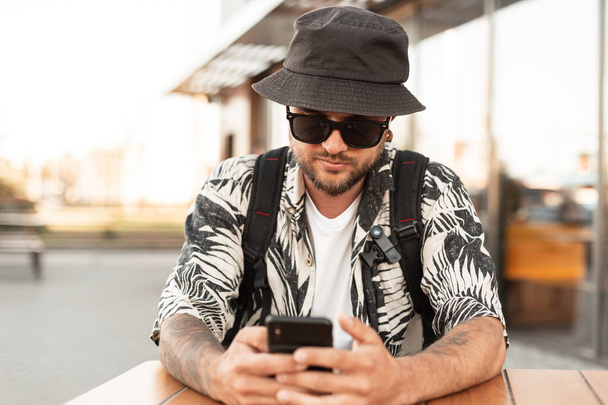 Handsome young hipster man in fashionable clothes with a hat sits in a summer cafe and communicates, surfs on a smartphone. Guy with phone travels in the city - Foto, Imagem