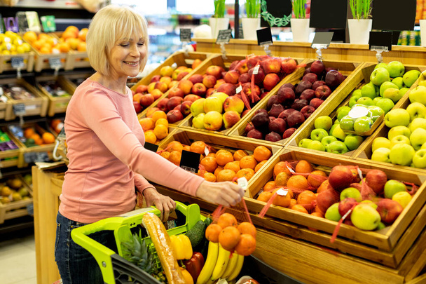 Vieille dame positive faisant du shopping dans un énorme supermarché, achetant des fruits - Photo, image
