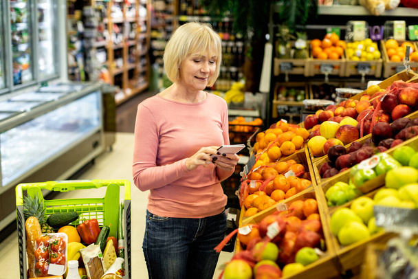 Atractiva anciana eligiendo frutas en el supermercado, sujetando el bloc de notas - Foto, imagen