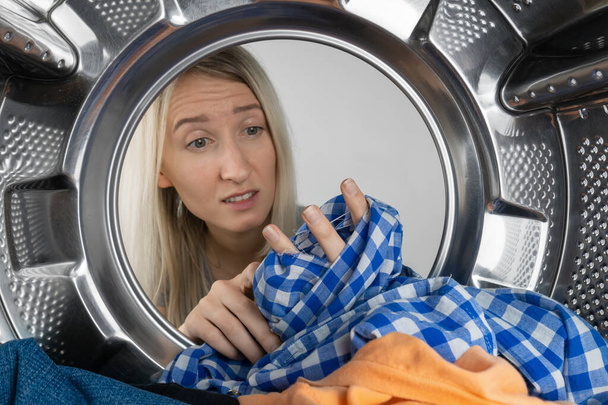 Disgruntled young blonde woman holds a leaky shirt in her hands after washing, photo from inside the drum of a washing machine. Woman is disappointed with the work of the washing machine - Фото, изображение