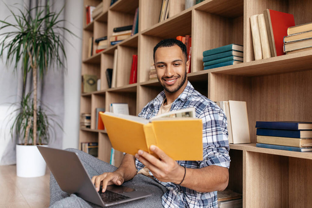 Happy arab man reading book near laptop computer, learning online while sitting at home - Φωτογραφία, εικόνα