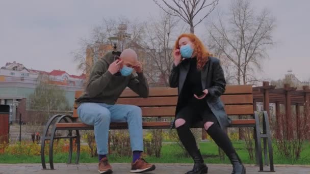 Man and woman sitting on park bench take off their protective masks and begin to communicate.  - Video