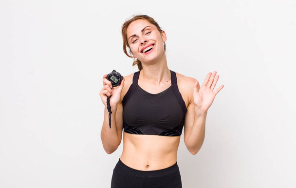 young pretty woman  smiling happily, waving hand, welcoming and greeting you. fitness concept and stopwatch - Foto, Bild