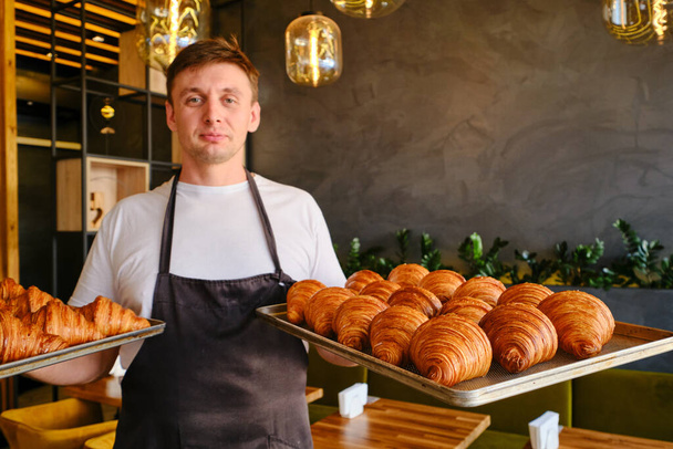 El hombre panadero sostiene dos bandejas para hornear de cruasanes dorados frescos en la panadería. Espacio para texto - Foto, imagen