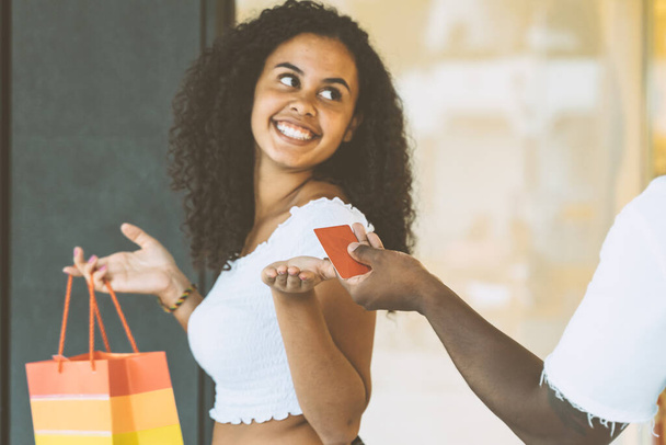 Молода африканська дівчина посміхається з кредитною карткою і сумками. Шопінг у продажу концепція свята сезону. Жінка в торговому центрі на відкритому повітрі протягом дня
. - Фото, зображення