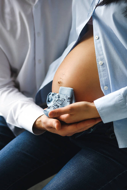妊娠中の女性と彼女の夫の写真を閉じます妊娠中の腹の屋内の近くに青いベビーソックスを保持.家族の概念。幸せな母と親の概念 - 写真・画像