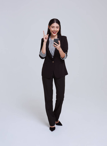 красивое и дружелюбное лицо азиатская деловая женщина улыбается в официальном костюме держа мобильный телефон на сером фоне. - Фото, изображение