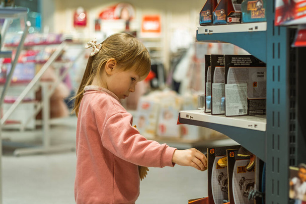 Klein blank meisje dat een nieuw speeltje kiest in de grote babywinkel. Grote planken vol speelgoed - Foto, afbeelding