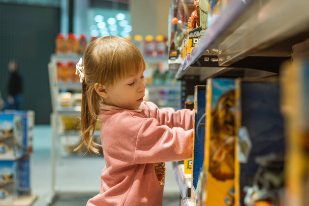 Маленькая белая девочка выбирает новую игрушку в большом детском магазине. Большие полки полные игрушек - Фото, изображение