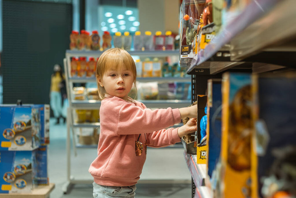 Küçük beyaz kız büyük bebek mağazasında yeni bir oyuncak seçiyor. Oyuncak dolu büyük raflar - Fotoğraf, Görsel