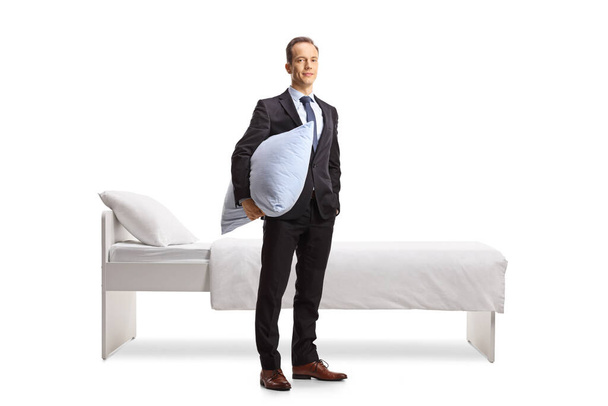 Businessman holding a pillow and looking at camera in front of a bed isolated on white background - Zdjęcie, obraz