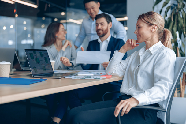 Usmívající se sebevědomá žena šéf sedí na setkání a dívá se na straně se svými kolegy v pozadí v kanceláři - Fotografie, Obrázek