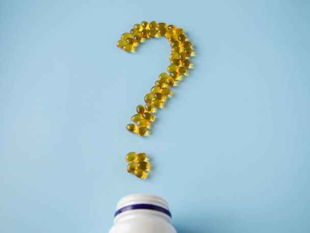 Vitamiinikapselit sinisellä pinnalla kysymysmerkkinä. Omega ja D-vitamiini Terveydenhuolto. Ravintolisät. Avattu pullo, jossa on keltaisia pillereitä, tabletteja ja kapseleita. Lääketieteelliset kysymykset. - Valokuva, kuva