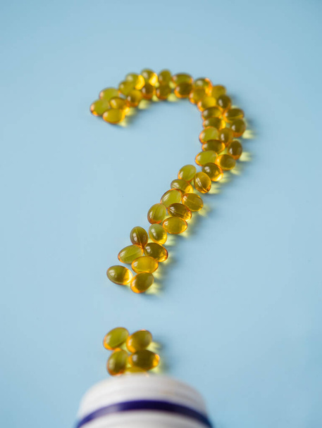 Vitamin capsules on a blue surface in the form of a question mark. Omega and vitamin D. Health care. Nutritional supplements. Opened bottle with yellow pills, tablets and capsules. Medicine questions. - Fotografie, Obrázek