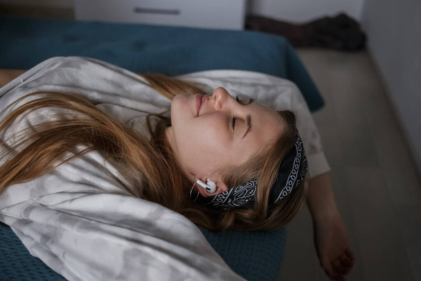 Kafasında bandajla ve uzun sarı saçlarıyla genç bir kız yatağa uzanıp kulaklıklarıyla müzik dinliyor. - Fotoğraf, Görsel