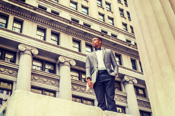 Wearing gray blazer, bow tie, black pants, holding laptop computer, a young black man standing on street. confidently looking up. Instagram effect - Photo, Image