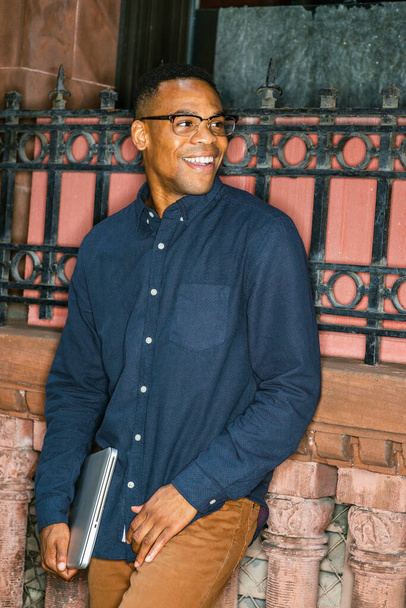 Mavi gömlekli, kahverengi pantolonlu, gözlüklü, dizüstü bilgisayarlı, genç adam caddede pencerenin önünde duruyor, gülümsüyor, - Fotoğraf, Görsel