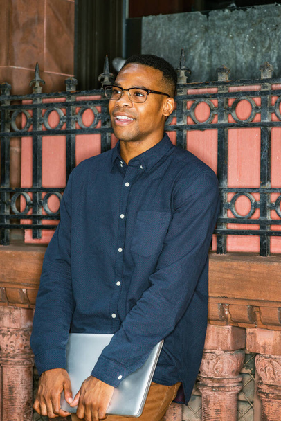 Kék inget visel, barna nadrágot, szemüveget, laptopot tart, egy fiatal fekete férfi áll az ablaknál az utcán, mosolyog, vár magára. - Fotó, kép