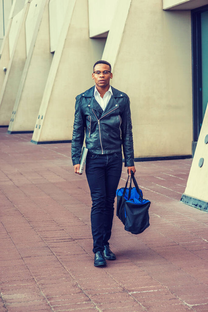 Wearing black leather jacket, jeans, leather shoes, glasses, holding laptop computer, carrying duffel bag, an African American college student walking on street in New York. - Photo, Image