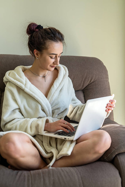 mooi jong meisje in een sneeuwwitte badjas met een laptop op haar knieën maakt online winkelen. Online winkelen concept. - Foto, afbeelding