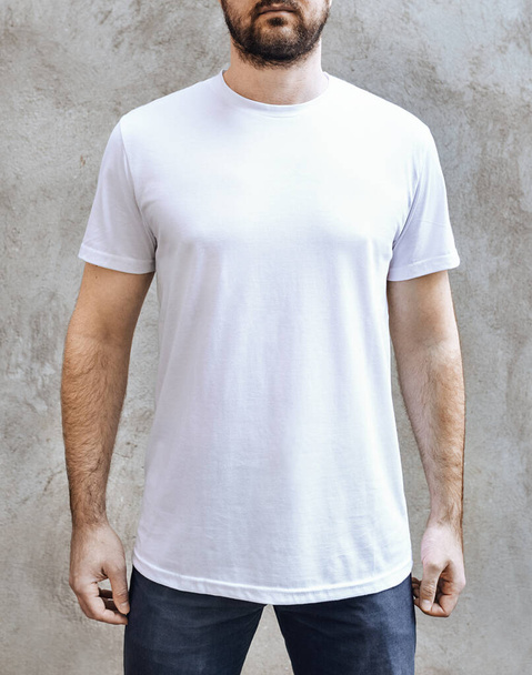 Young man with a beard in a blank white casual t-shirt. Front view on a background of light gray concrete wall. Design and layout of men's t-shirt for printing. - Photo, Image