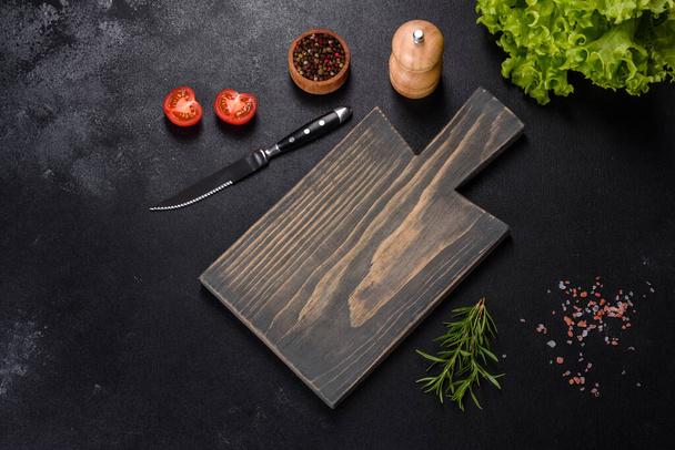 A wooden cutting board with a kitchen knife with spices and herbs on a dark concrete background. Cooking at home - Foto, Bild