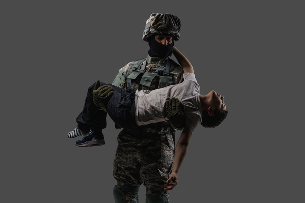 Γενναίος πατριώτης στρατιώτης που κουβαλάει τραυματισμένο παιδί σε γκρι φόντο - Φωτογραφία, εικόνα